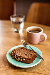 A slice of banana bread with pumpkin seeds on a blue plate, a dessert fork, a pink cup of hot black coffee, a glass of water. Wooden table, cafe.