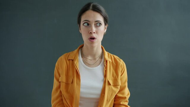 Slow motion portrait of confused young lady rolling eyes then looking at camera with shy face standing on gray background. People and expressions concept.