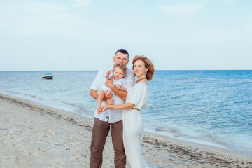 A young family with a little daughter, a toddler, in light clothes in the summer on the seashore, enjoy their vacation. Happy family and children. Summer vacation and tourism