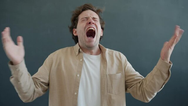 Slow motion portrait of stressed young man screaming and flapping arms expressing negativity then looking at camera on gray background. Emotions and stress concept.
