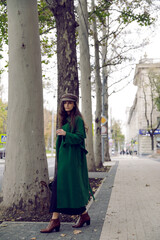 Elegant young woman , dressed in a long green coat and hat, posing in the street, alley background.