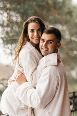 Young beautiful happy newlyweds in love a man and a woman in white bath robes on the balcony of the house smile and hug