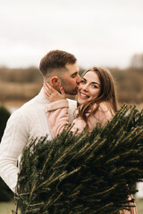 Young beautiful happy lovers man and woman, love story in winter with a live Christmas tree in their hands on the background of the river bank