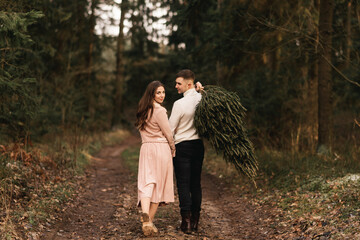 Young beautiful happy lovers man and woman, love story in winter with a live Christmas tree in a pine forest in the sun