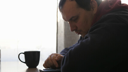 Handsome man at restaurant using smartphone. Man using smartphone touchscreen in cafe by window. Handsome man using digital display touch screen device in cafe and drinks coffee.