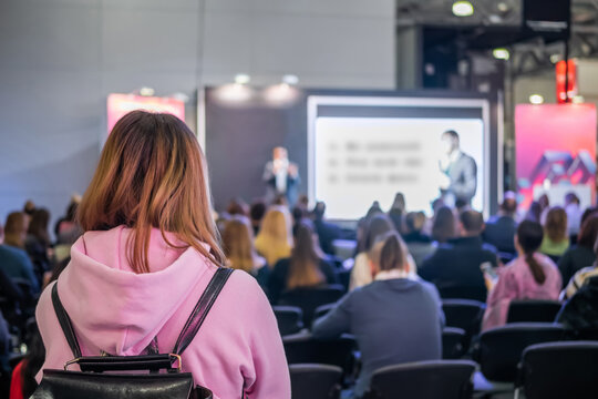 Business, Congress, Seminar, Corporate, Training, Education Concept. Back View Of Unrecognizable Woman In Pink Hoody Listening Lecture, Presentation At Trade Show, Conference Hall