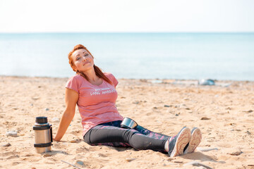 Fototapeta na wymiar An adult woman in sportswear, sitting relaxed on the sand, next to a shaker. In the background, the ocean and sky. Copy space. Sporty lifestyle concept