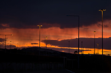 Fototapeta na wymiar Spectacular red sunset with dark rainy clouds over the bridge with silhouettes of people and vehicles and tall street lanterns along highway M50 in Dublin, Ireland. Beautiful cold sunset clouds