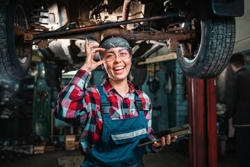 Fototapeta na wymiar A young beautiful happy female mechanic, in a uniform and glasses, with a tablet in her hands, poses standing under a car on a lift