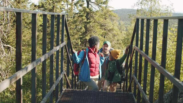 Multiethnic Family Walking With Backpacks On Trail In National Park, Looking At Nature Around And Discussing Scenic View While Hiking Together On Summer Vacation