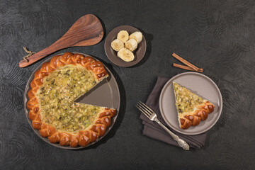 A banana pie with a sliced piece on a flat plate with a fork on a dark background.