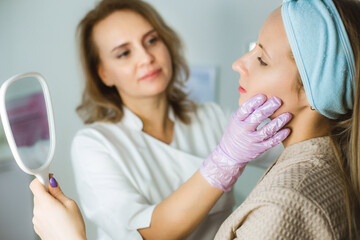 Facial skin care and protection. A young woman at a beauticians appointment looks in a mirror. A specialist examines the skin