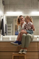 Vertical full length portrait of young mother helping little girl with down syndrome brush teeth in...