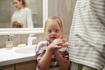 Portrait of cute girl with down syndrome looking at camera while standing in bathroom with mother,...