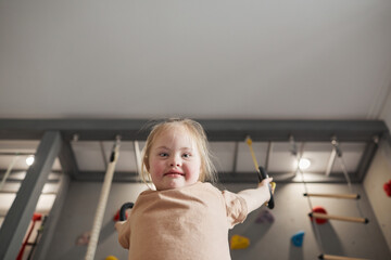 Low angle portrait of cute girl with down syndrome swinging on rings and enjoying exercise at home or at sports center while looking at camera, copy space
