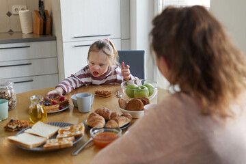 Portrait of cute girl with down syndrome reaching for sweet waffles while enjoying breakfast with family at home, copy space