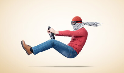 A man in stylish goggles with a scarf on his face flies forward holding a car steering wheel in his...