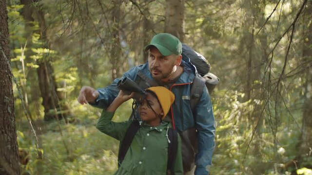 Tilt down shot of Caucasian father talking and showing something to little mixed race son as he looking through binoculars in woods