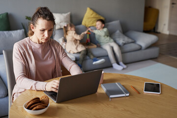 Portrait of single mother using laptop while working from home with two children playing in...