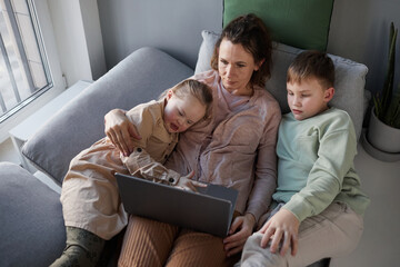 Top down view at caring mother lying on couch embracing son and daughter with down syndrome while watching videos via laptop, copy space