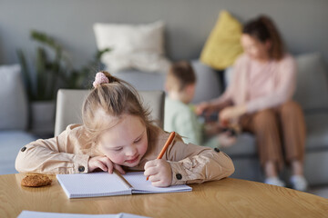 Portrait of cute girl with down syndrome doing homework at table in living room with mom and brother playing in background, copy space