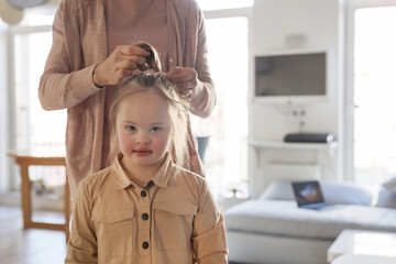 Cropped shot of caring mother tying hair of daughter with down syndrome in cozy home interior, copy...