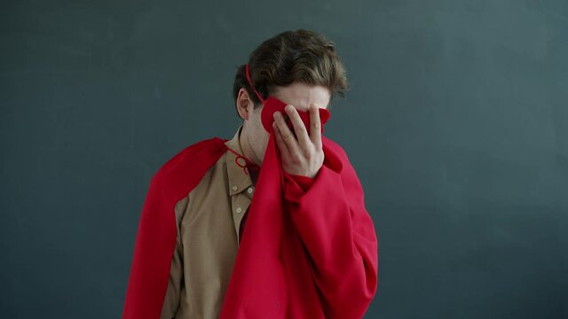 Portrait of unhappy guy wearing super hero costume crying and wiping tears with red cape standing on gray background expressing negative emotions.