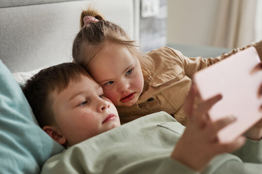 Portrait of cute girl with down syndrome using digital tablet with brother while lying in bed with brother, copy space - Powered by Adobe