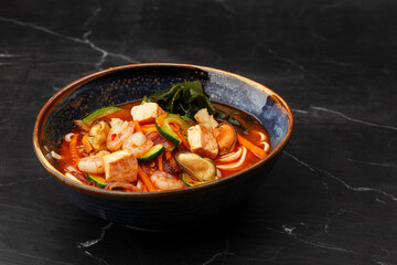 Asian ramen soup with shrimps, mussels, tofu, carrot, zucchini and seaweed. Dish isolated in a dark blue bowl, close-up on a black marble background. Asian cuisine.