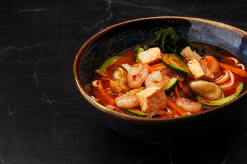 Asian ramen soup with shrimps, mussels, tofu, carrot, zucchini and seaweed. Dish isolated in a dark blue bowl, close-up on a black marble background. Asian cuisine. Copy space.