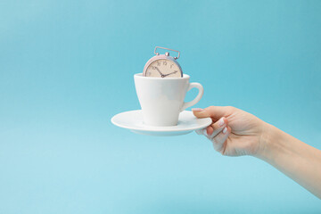 An old clock in a white cup on a plate held by a woman's hand on a pastel blue background.