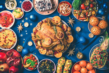 New year dinner table. Roasted Christmas chicken with orange slices, cranberries, garlic, festive decoration, candles, tangerine, pomegranate, golden glitter stars on blue background
