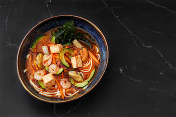 Asian ramen soup with shrimps, mussels, tofu, carrot, zucchini and seaweed. Dish isolated in a dark blue bowl, close-up on a black marble background. Asian cuisine. Copy space, top view.