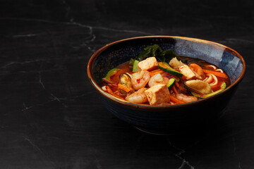 Asian ramen soup with shrimps, mussels, tofu, carrot, zucchini and seaweed. Dish isolated in a dark blue bowl, close-up on a black marble background. Asian cuisine. Copy space.