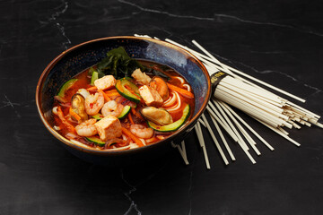 Asian ramen soup with shrimps, mussels, tofu, carrot, zucchini and seaweed. Dish served in a dark blue bowl, isolated on a black marble background with dry noodles by side. Asian cuisine.