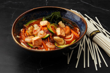 Asian ramen soup with shrimps, mussels, tofu, carrot, zucchini and seaweed. Dish served in a dark blue bowl, isolated on a black marble background with dry noodles by side. Asian cuisine.