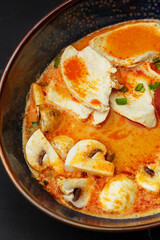 Asian chicken curry soup with chicken breast, tomatoes, mushrooms and spring onion. Close-up photo of the dish served in a dark blue bowl, isolated on a black marble background. Asian cuisine.