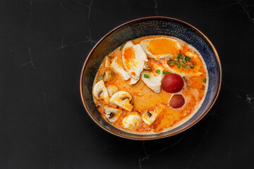 Asian chicken curry soup with chicken breast, tomatoes, mushrooms and spring onion. Close-up photo of the dish served in a dark blue bowl, isolated on a black marble background. Asian cuisine.