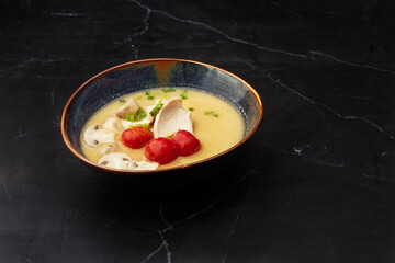 Asian coconut chicken soup with chicken breast, tomatoes, mushrooms and spring onion. Close-up photo of the dish served in a dark blue bowl, isolated on a black marble background. Asian cuisine.