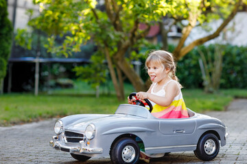 Fototapeta na wymiar Little adorable toddler girl driving big vintage toy car and having fun with playing outdoors. Gorgeous happy healthy child enjoying warm summer day. Smiling stunning kid playing in domestic garden