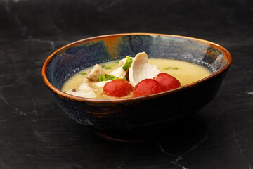 Asian coconut chicken soup with chicken breast, tomatoes, mushrooms and spring onion. Close-up photo of the dish served in a dark blue bowl, isolated on a black marble background. Asian cuisine.