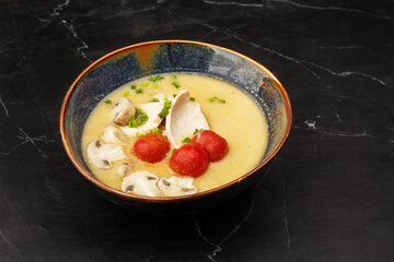 Asian coconut chicken soup with chicken breast, tomatoes, mushrooms and spring onion. Close-up photo of the dish served in a dark blue bowl, isolated on a black marble background. Asian cuisine.