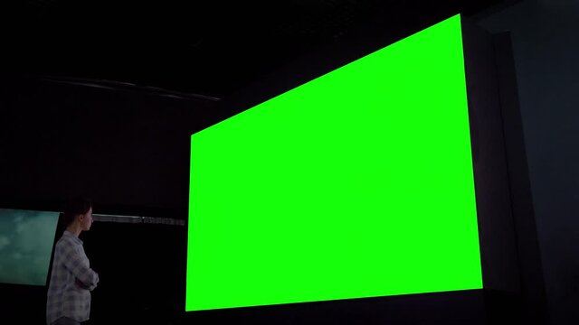 Green screen, technology, chroma key, futuristic, template, mock up, entertainment concept. Woman looking at large wall blank interactive green display in dark room of science exhibition or museum