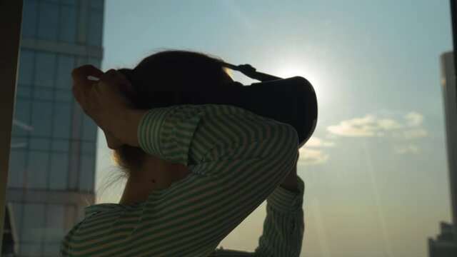 Slow motion: woman using virtual reality headset and looking around against view on cityscape from panoramic skyscraper window in office. Sun lens flare. VR, sightseeing, discover, technology concept