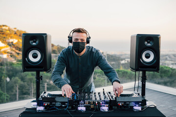 Caucasian man with headphones and surgical mask, playing music on a DJ table with big speakers,...