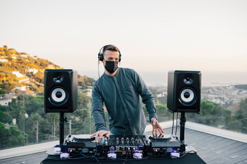 Professional Techno male DJ in surgical mask and headphones playing music at a home terrace during...