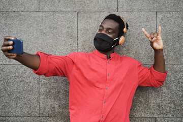 African young man listening music on mobile phone app while wearing protective mask - Focus on face