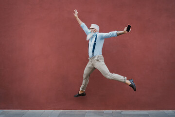 Senior crazy man jumping outdoors while wearing mask with red wall in background - Focus on face