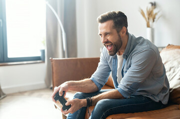 Portrait of young bearded hipster guy sitting on the couch in the living room, holding joystick,...
