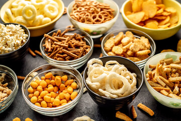 Assorted chips, popcorn, onion rings, croutons, cheese balls, biscuits in bowls. unhealthy food for figure, heart, skin, teeth. An assortment of fast carbohydrates. Junk food on a stone background